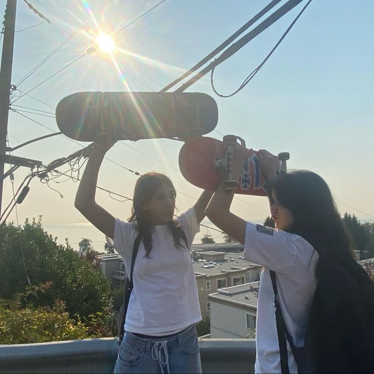 two girls holding up skateboards on top of a roof in front of the sun