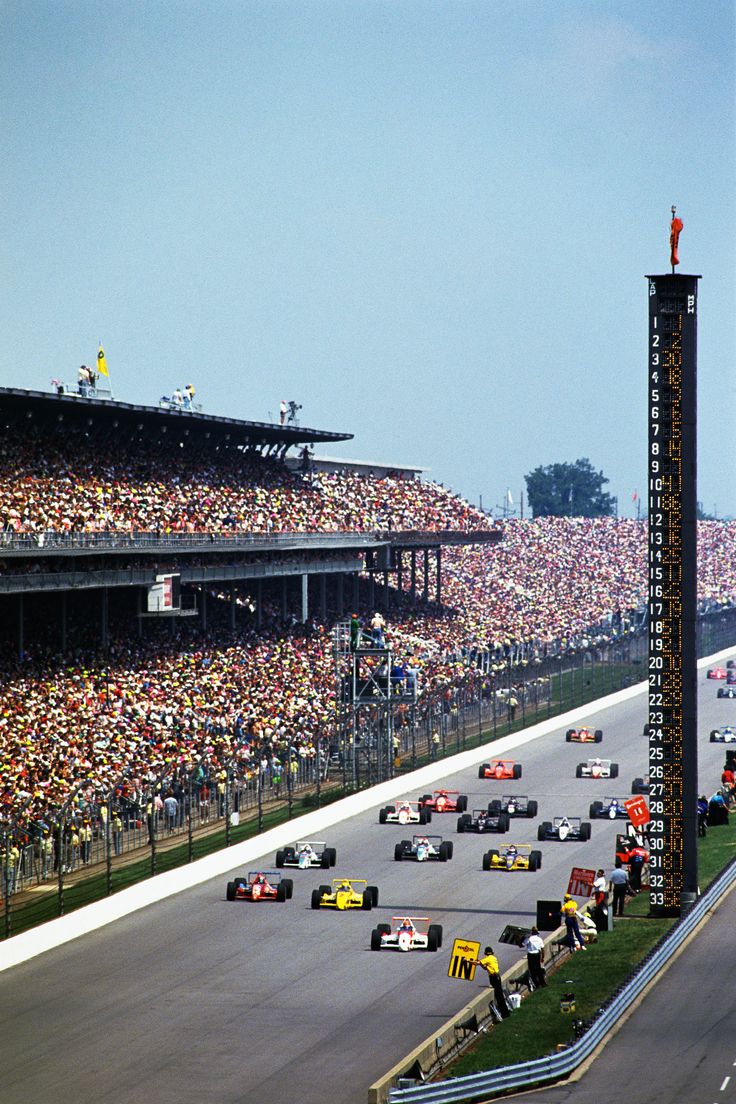 a group of cars driving down a race track in front of a crowd of people