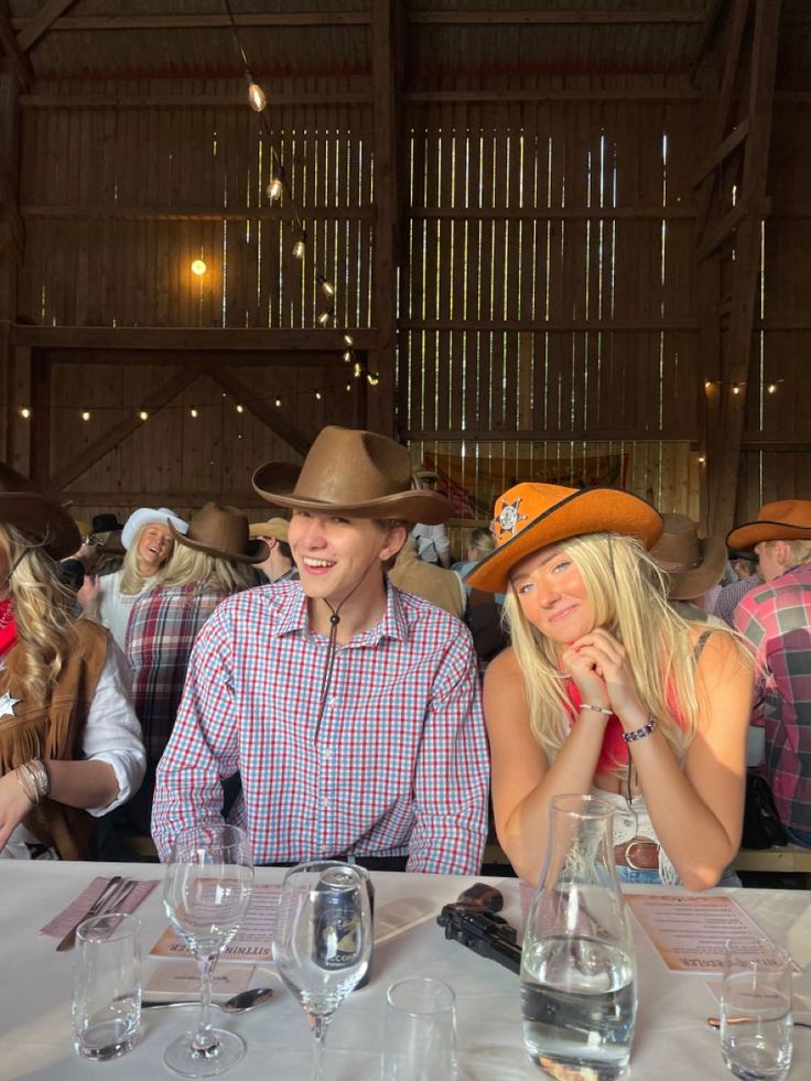 two people sitting at a table with wine glasses in front of them and one person wearing a cowboy hat