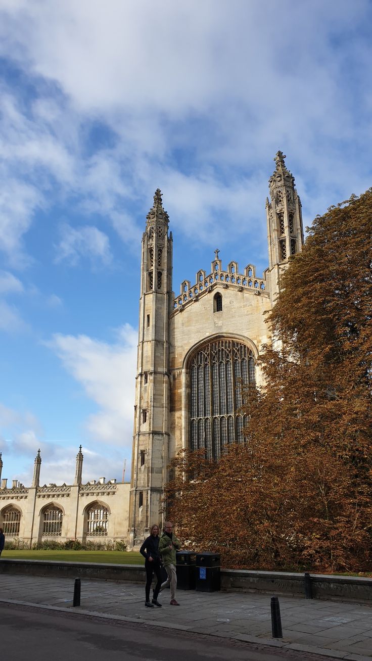 two people walking in front of a large building with tall towers and windows on it