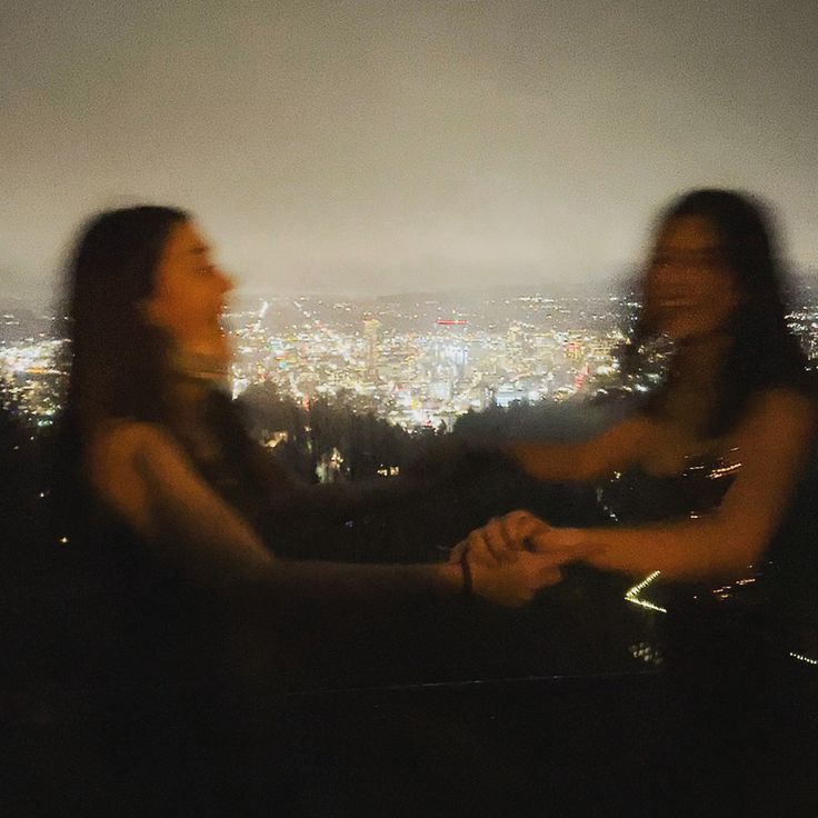 two women standing next to each other in front of a city at night with lights on