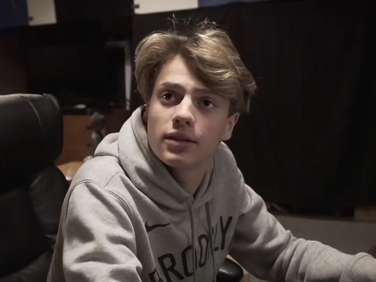 a young man sitting in front of a computer monitor wearing a hoodie and looking at the camera