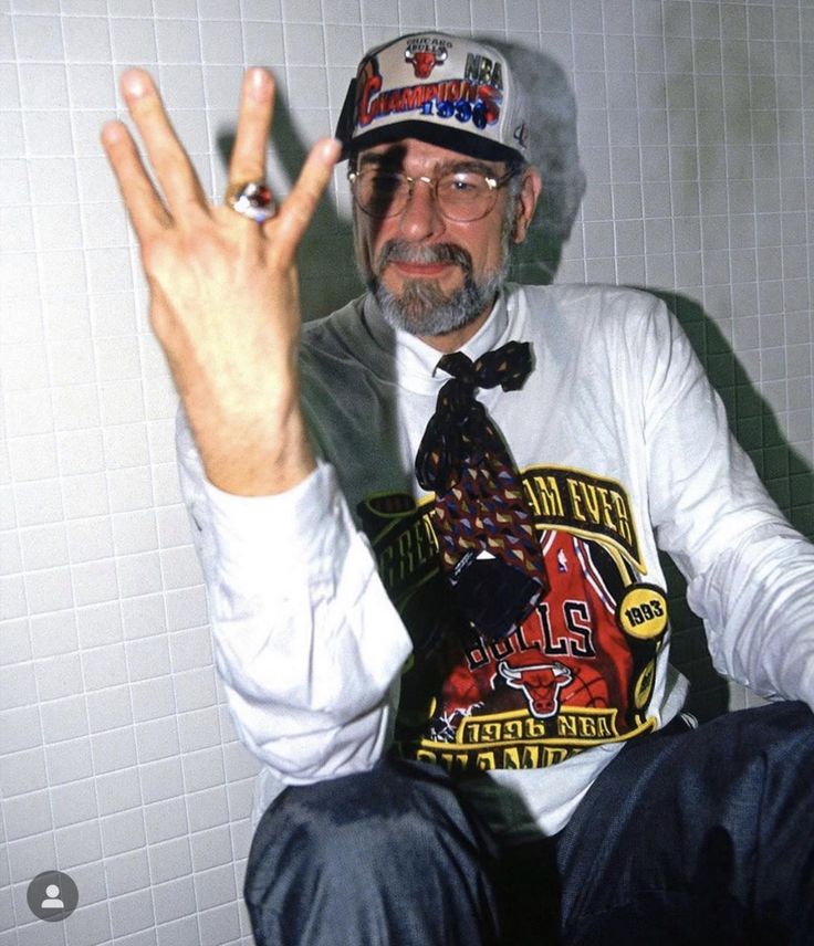 an older man wearing a hat and tie sitting on a toilet with his hand up in the air