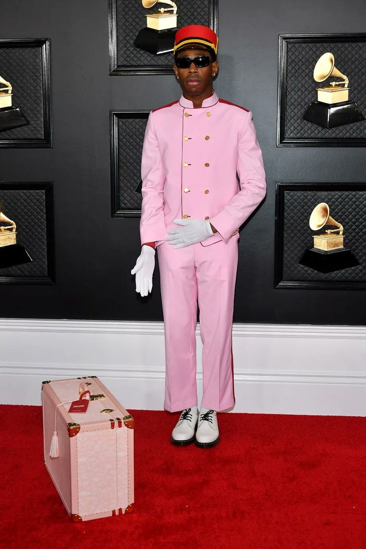 a man in a pink suit and hat standing next to a suitcase on a red carpet