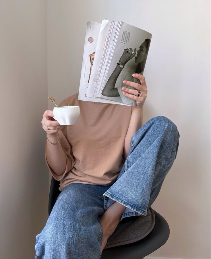 a woman sitting in a chair reading a magazine and holding a cup with her hand