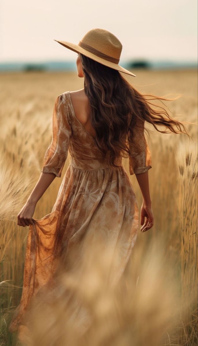 a woman in a dress and hat walking through a wheat field with her hair blowing in the wind