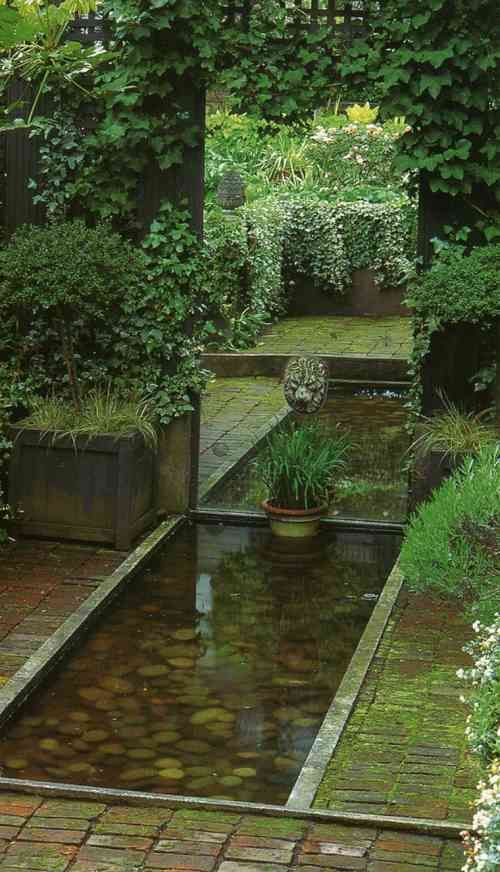 a small pond surrounded by greenery and brick walkways in the middle of a garden