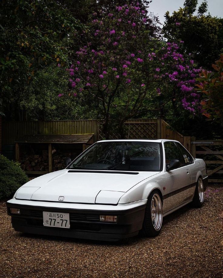 a white car is parked in front of some trees and purple flowers on a cloudy day