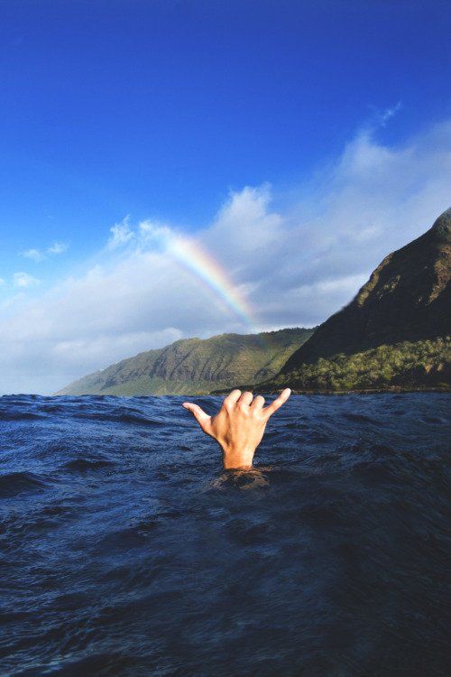 a hand reaching out from the water with a rainbow in the sky above it and mountains in the background