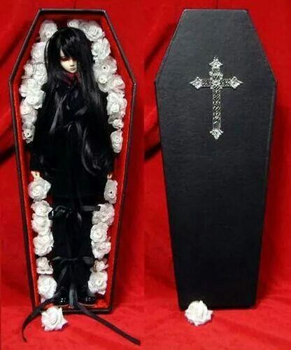 a doll in a black outfit standing next to a casket with roses around it and a cross on top