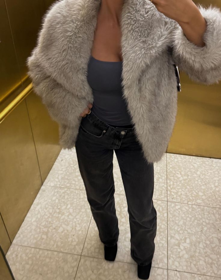 a woman taking a selfie in front of a mirror wearing a fur coat and jeans