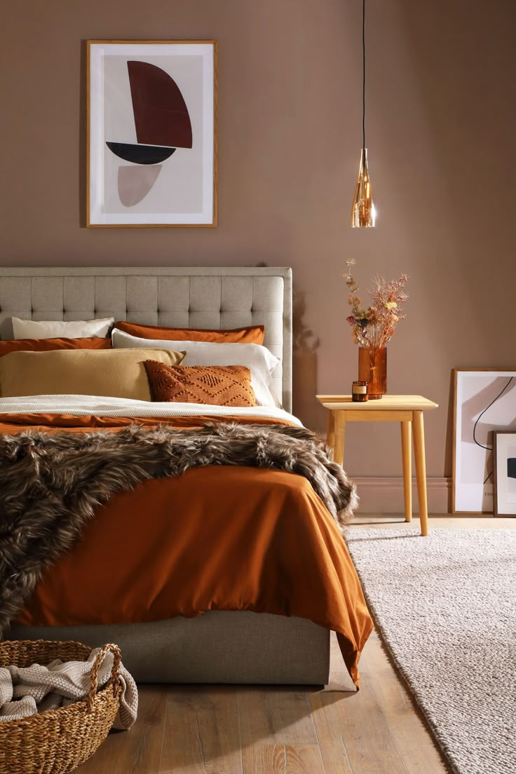 a bed with orange and brown sheets in a bedroom next to a table, lamp and pictures on the wall