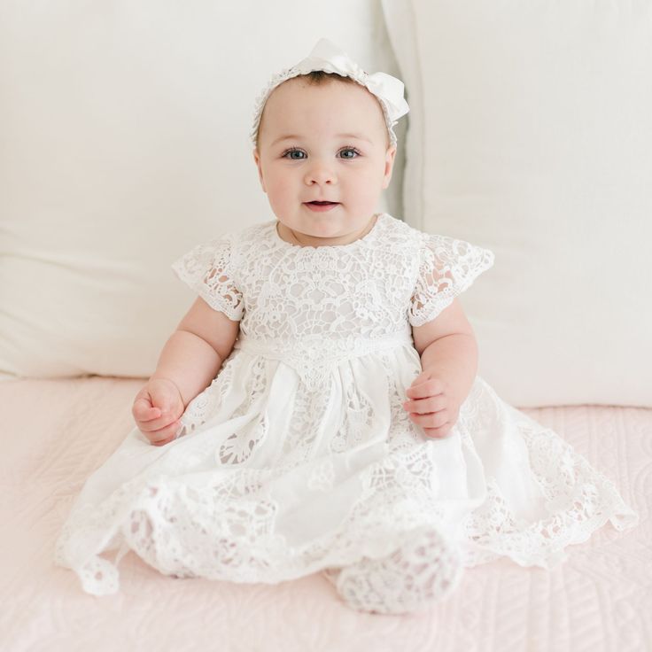 A baby wearing the Grace Dress, a white lace flower girl dress, and a matching headband with a bow sits on a light pink quilted surface, with white pillows in the background. The baby is smiling and has hands resting gently on the lap. First Birthday Family Photos, Birthday Family Photos, White Baby Dress, Her First Birthday, First Birthday Dress, Grace Dress, First Birthday Dresses, Grace And Lace, Baby Pics