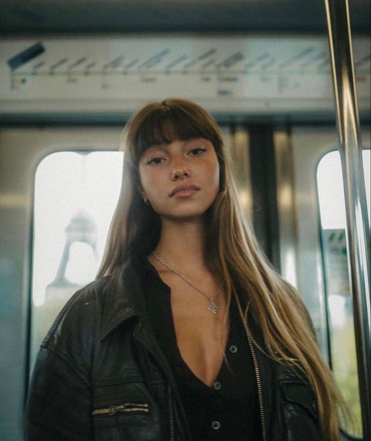 a woman with long hair standing in front of a window on a subway train, looking at the camera