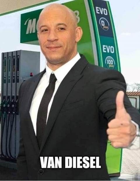 a bald man in a suit giving the thumbs up sign at an ev ev gas station