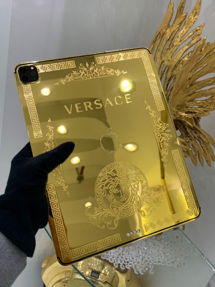 a person holding up a gold versa phone case in front of a clock and other items