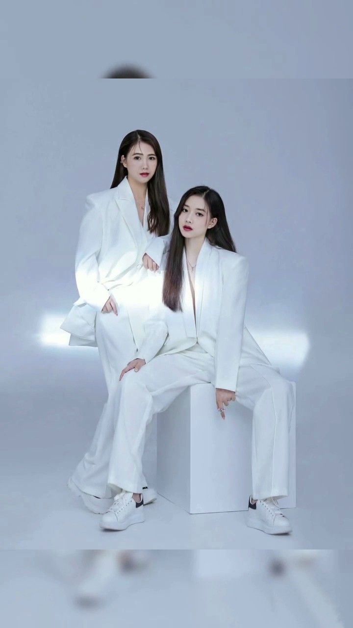 two young women sitting on top of a white bench in front of a gray background