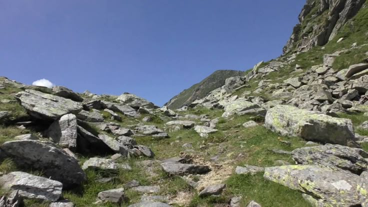 some rocks and grass on the side of a mountain with a blue sky in the background