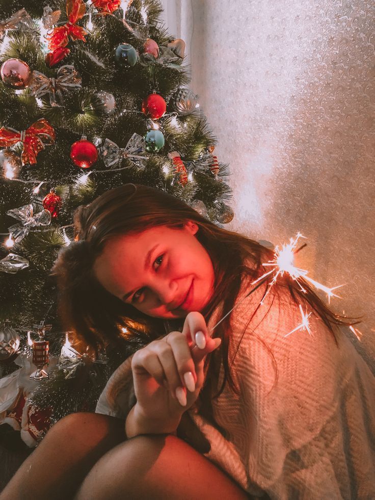 a woman sitting in front of a christmas tree with sparklers on her finger and looking at the camera