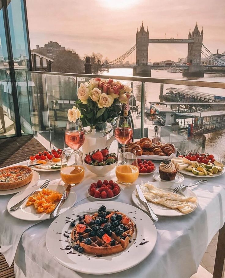 a table full of food and drinks on top of a balcony overlooking the water with a bridge in the background