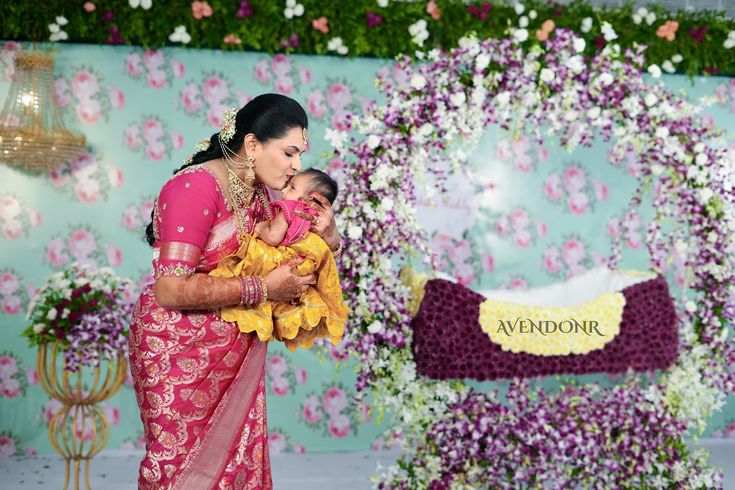 a woman holding a baby in her arms while standing next to a floral wall with flowers on it