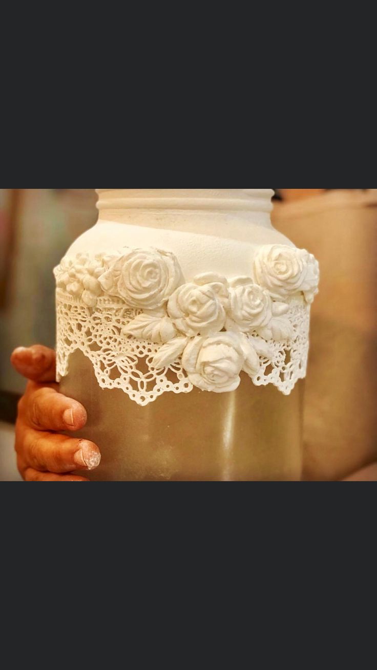 a person holding a vase with white flowers on the bottom and lace around it's edges