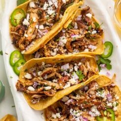 three tacos with shredded meat, onions and peppers on a white platter next to limes