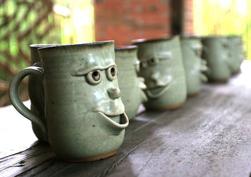 four green mugs with faces on them sitting on a wooden table outside in front of a brick wall