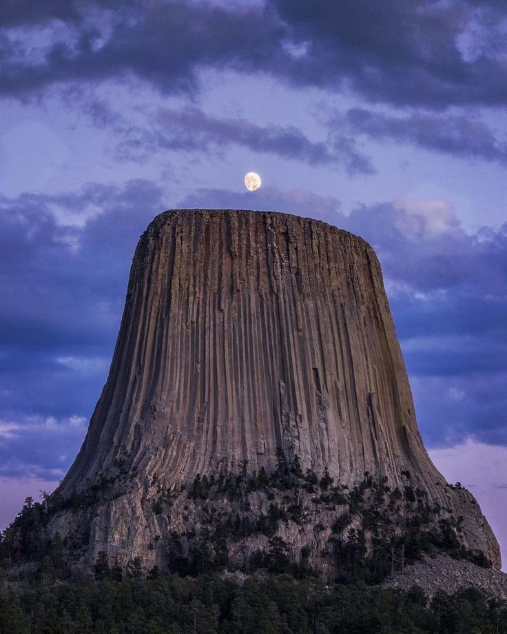 the moon is setting on top of a tall rock formation with trees in front of it