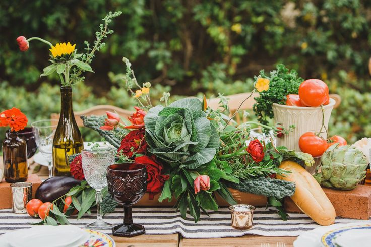 the table is set with flowers, vegetables and wine bottles for an outdoor dinner party