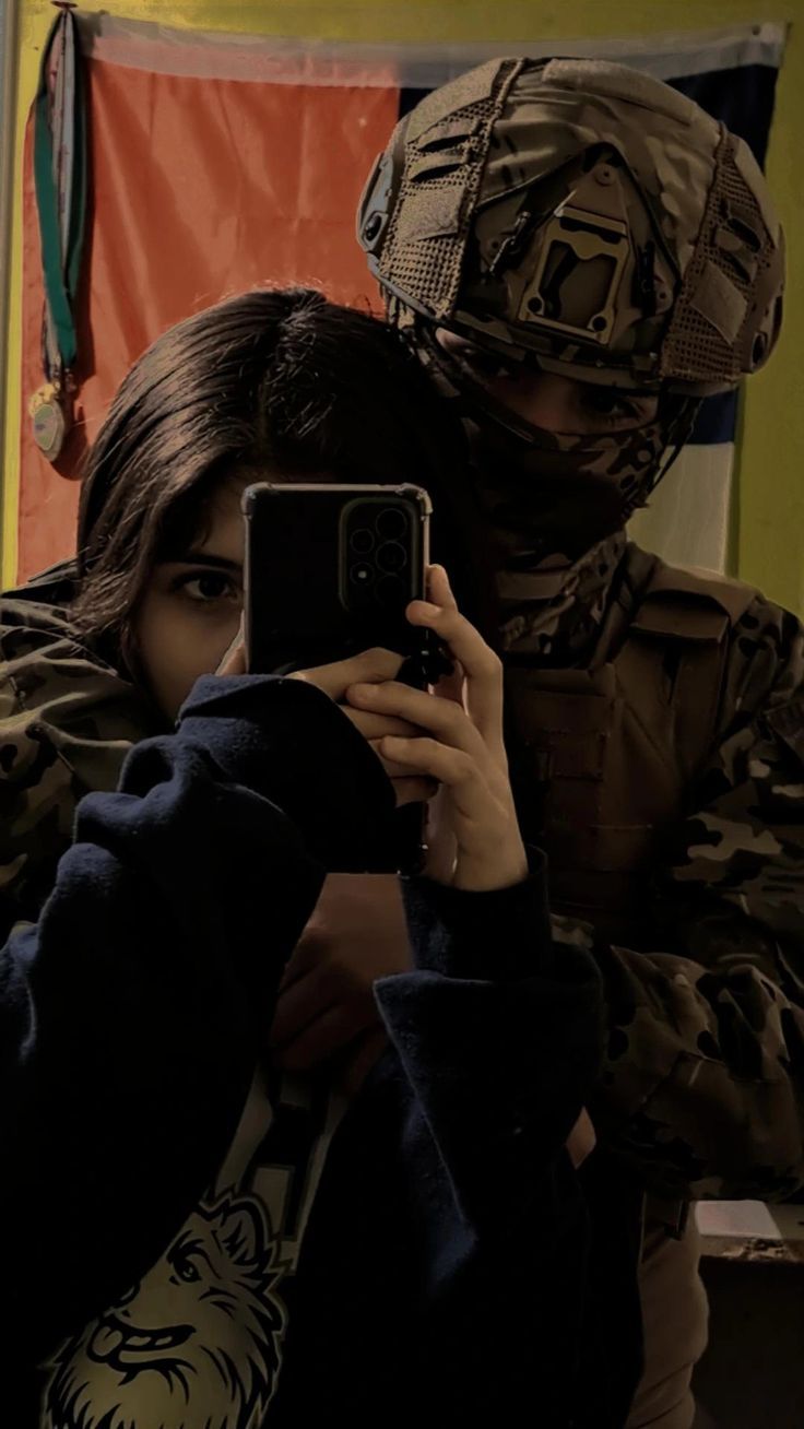 a woman taking a selfie in front of a soldier with her cell phone while wearing camouflage clothing