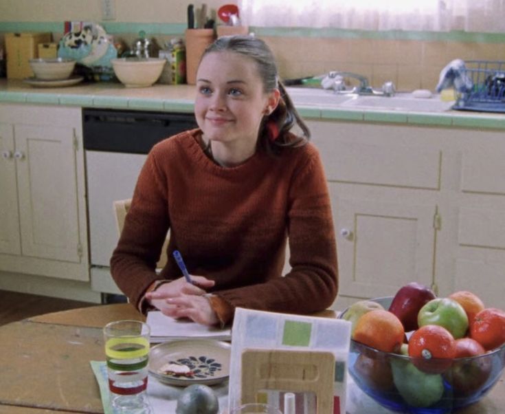 a woman sitting at a kitchen table in front of a bowl of fruit