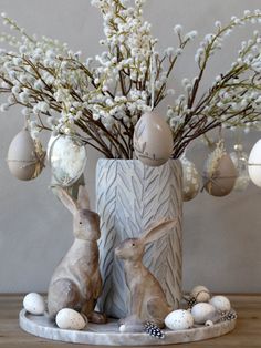 two bunny figurines sitting next to an egg vase with flowers and eggs in it