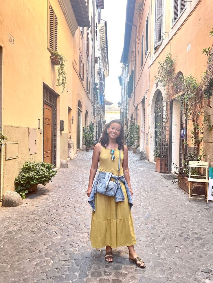 a woman standing in the middle of an alleyway wearing a yellow dress and carrying a blue purse