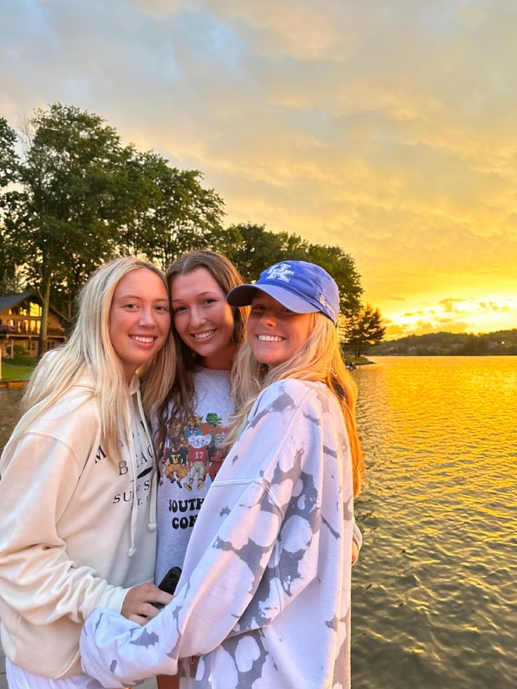 three girls are posing for the camera in front of a body of water at sunset