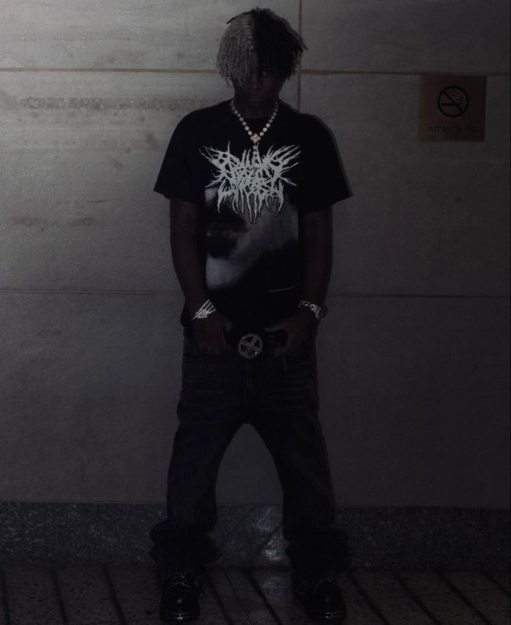 a young man standing in front of a wall wearing a black shirt and chain necklace