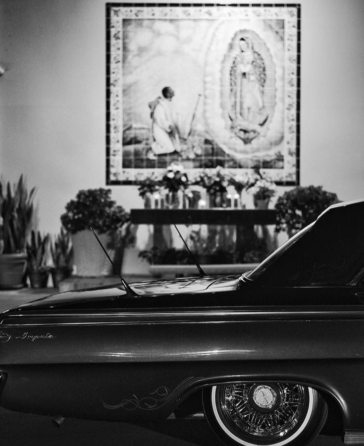 a black and white photo of an old car parked in front of a painting on the wall