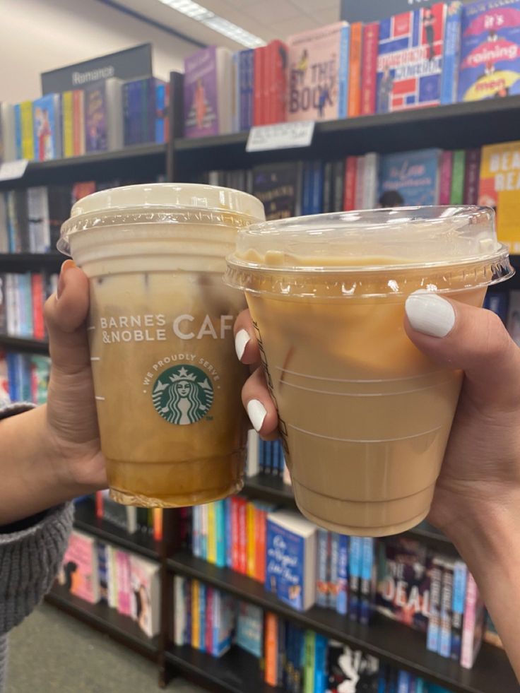 two people holding up starbucks cups in front of bookshelves