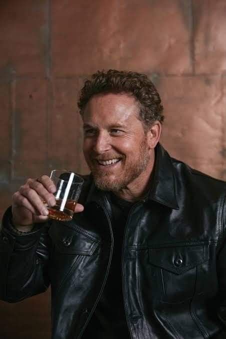 a man in a black leather jacket holding a glass with something in it's hand