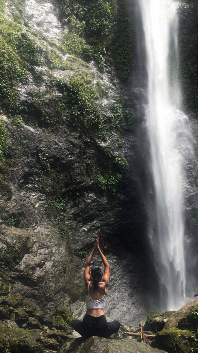 a woman sitting on rocks in front of a waterfall with her hands up to the sky