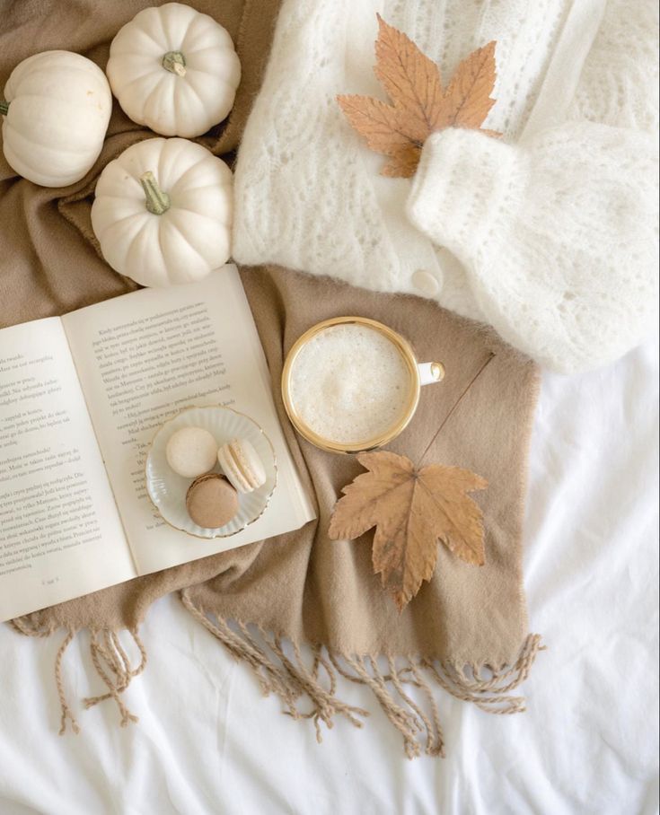 an open book and cup of coffee on a bed next to some autumn leaves, pumpkins and other items