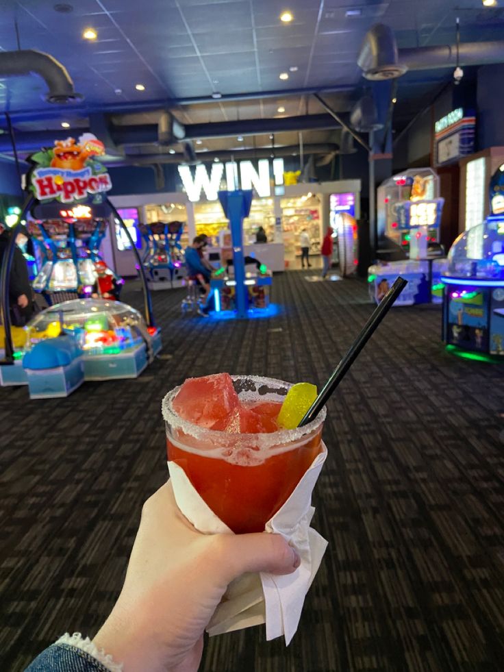 a person holding up a drink in front of a game room with arcade machines and other games