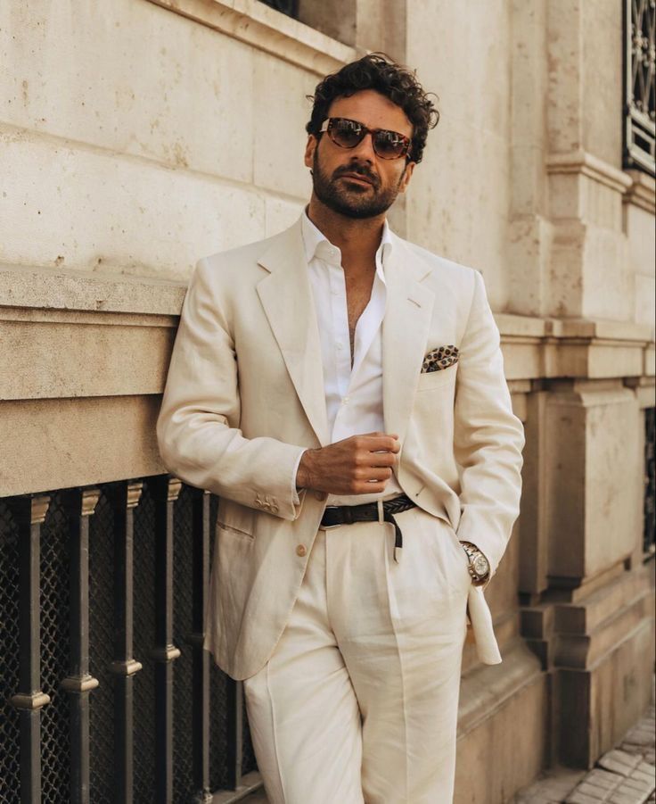 a man in a white suit leaning against a wall with his hand on his hip