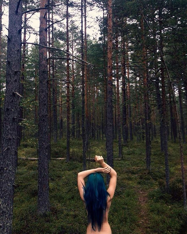 a woman with blue hair standing in the woods looking up at trees and holding an apple