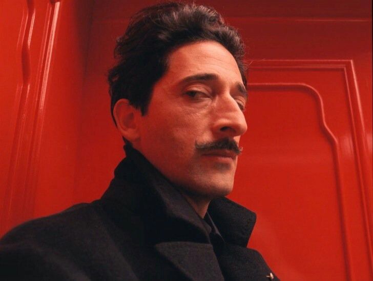 a man with a moustache standing in front of a red wall wearing a black coat