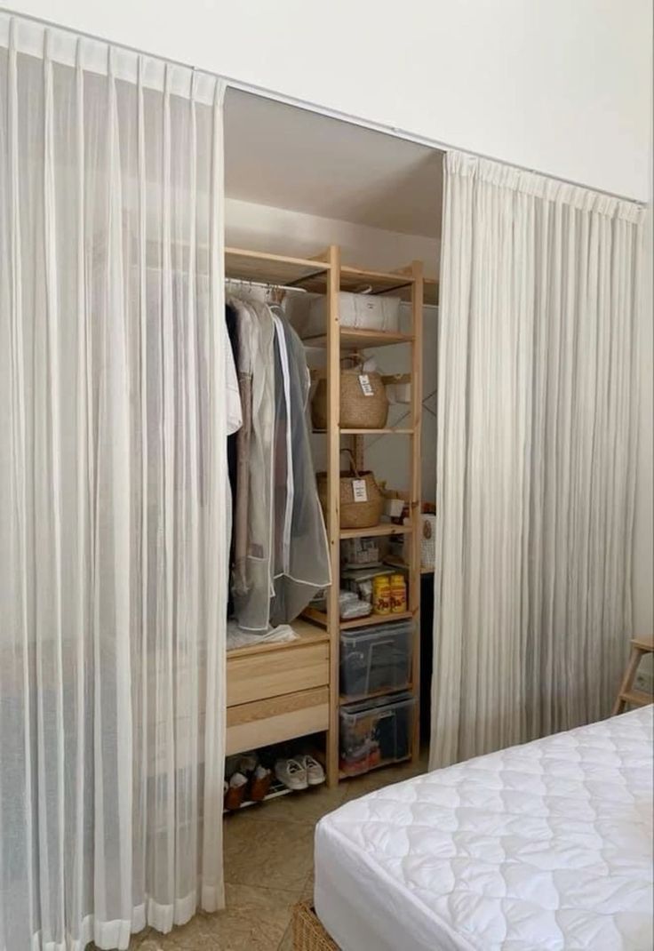 an open closet with white curtains and clothes on the shelves, next to a bed