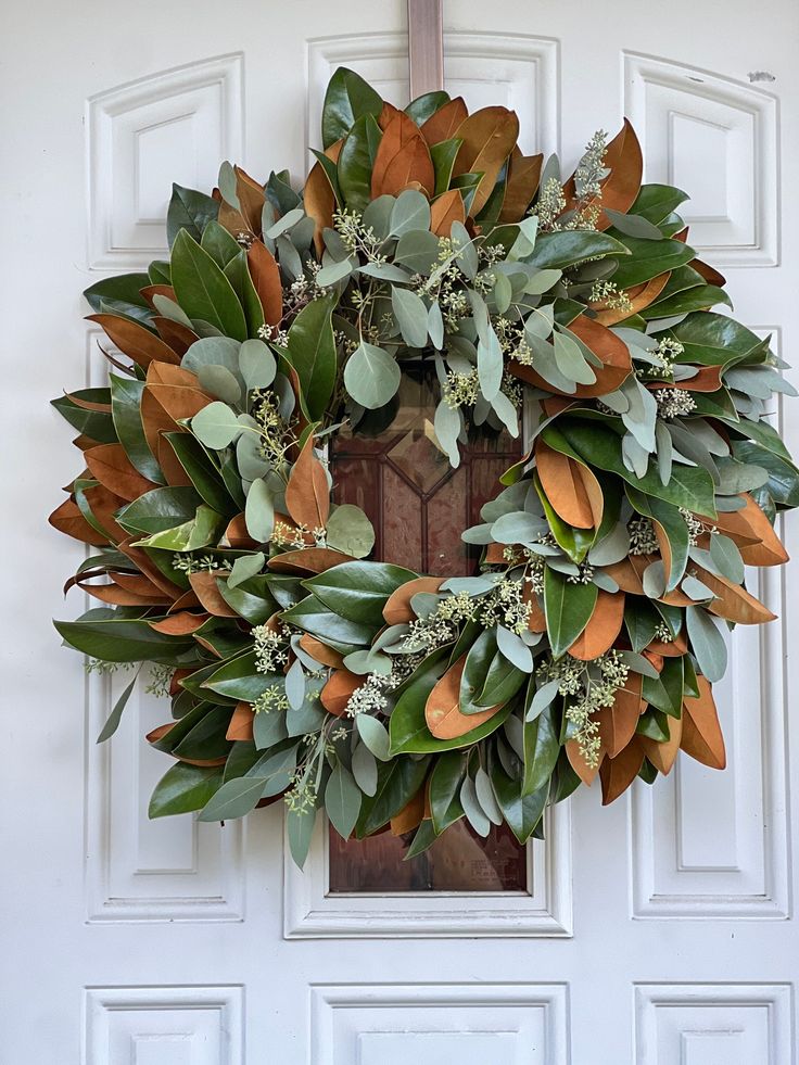 a wreath is hanging on the front door with leaves and greenery around it's edges