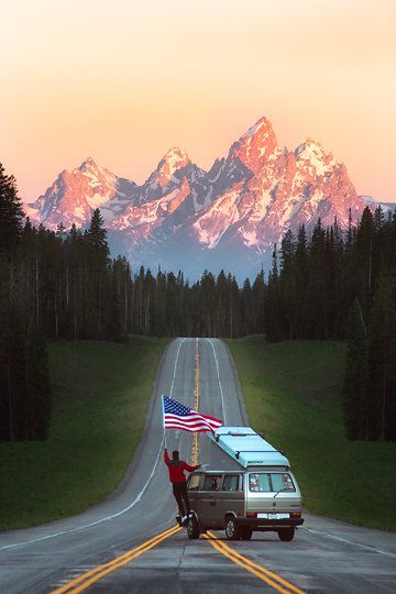 a van with an american flag on the back driving down a road in front of mountains