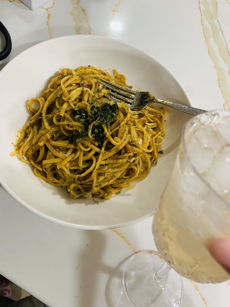 a white bowl filled with pasta and spinach on top of a table next to a glass of wine