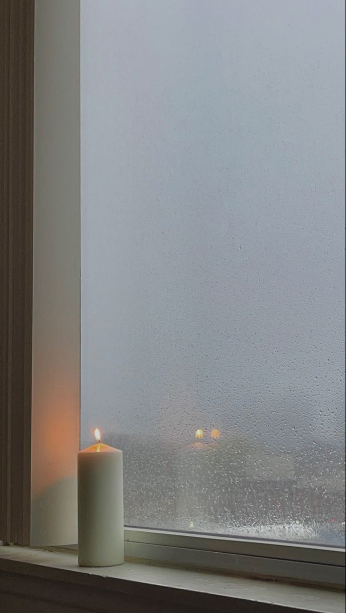 a white candle sitting on top of a window sill next to a rain covered window